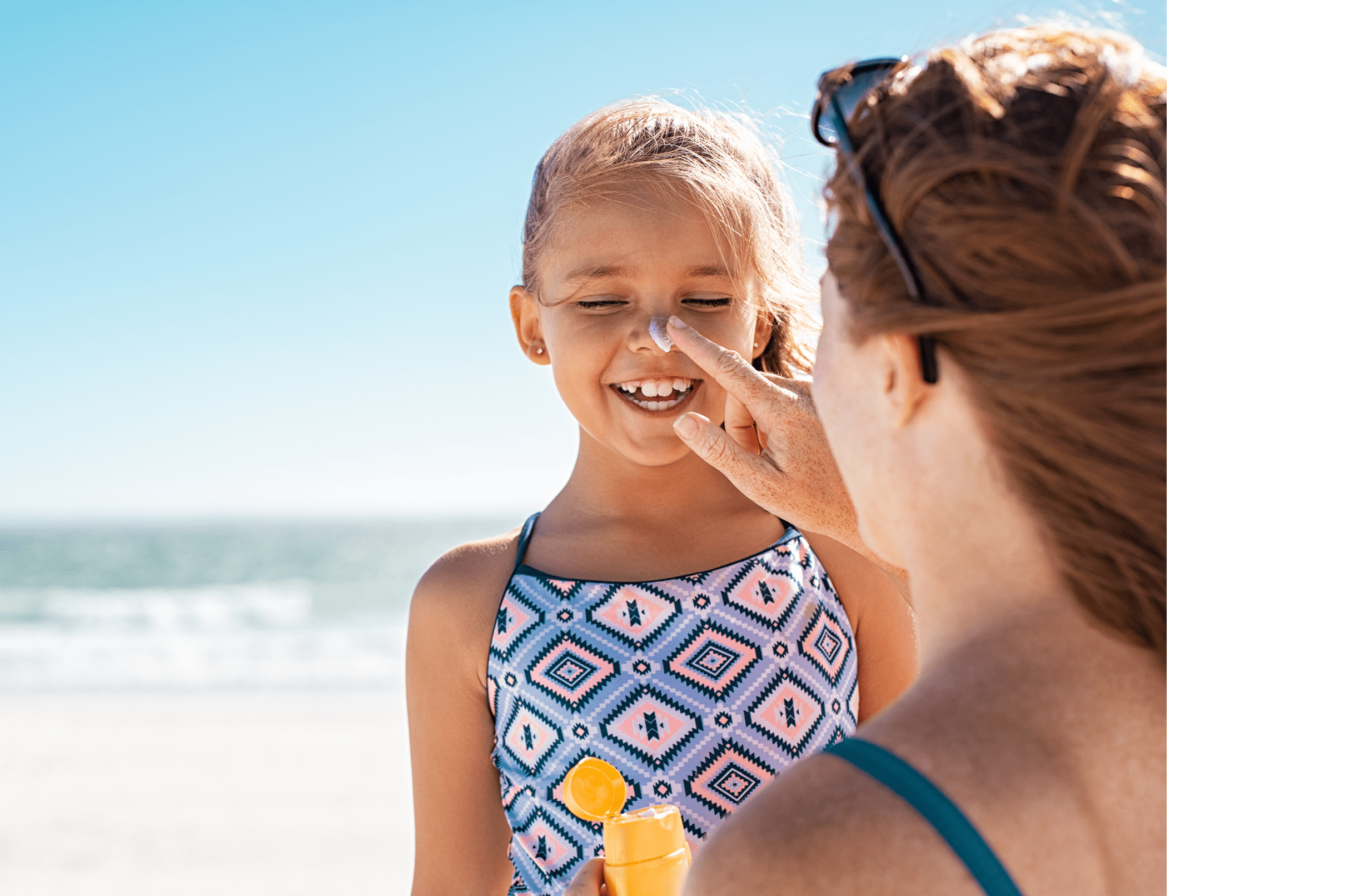 Young mother applying protective sunscreen on daughter nose at beach. Woman hand putting sun lotion on child face. Cute little girl with sunblock at seaside with copy space.