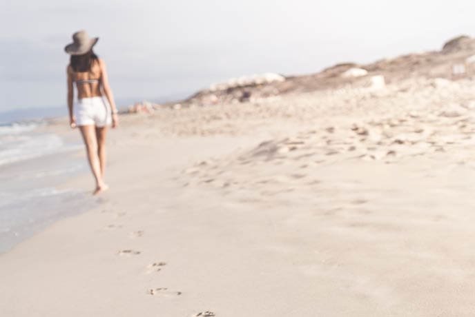 Distant Woman in shorts walking barefoot on white sandy beach in Formentera, Ibiza leaving footprints in sand. Focus is on the foreground footprints