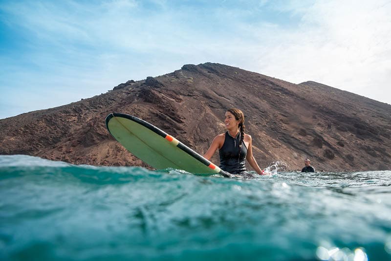 Beautiful young brunette woman surfing in Fuerteventura in the Atlantic ocean on the background of blue sky, clouds and transparent waves